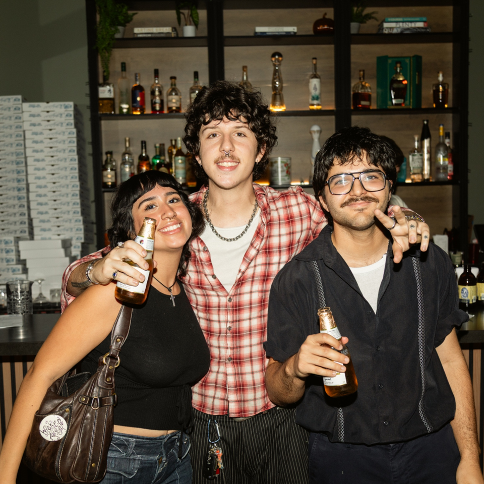 Three smiling friends pose together in a restaurant bar, holding bottled drinks before shelves stocked with liquor.