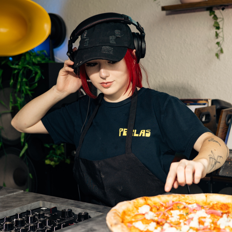 Red-haired person wearing headphones and an apron reaches toward a pizza beside audio equipment in a cozy studio.