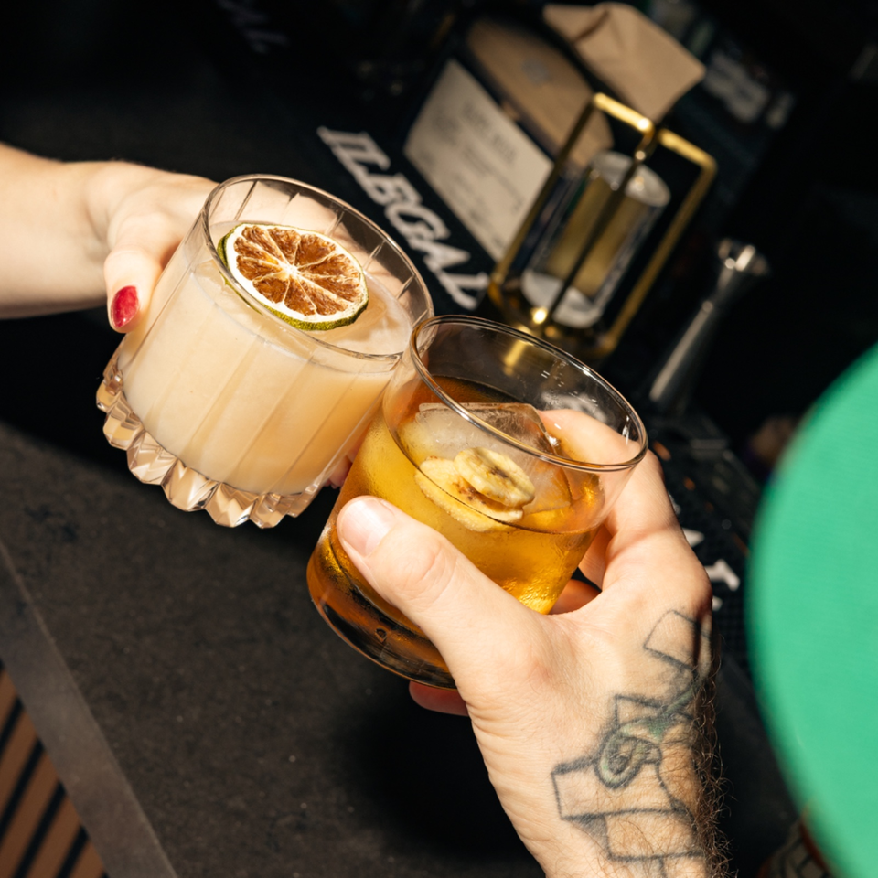 Two cocktails garnished with dried citrus slices are held over a dark restaurant table by tattooed hands.
