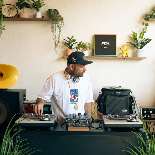A DJ in a hat and headphones plays records on turntables in a plant-filled room.