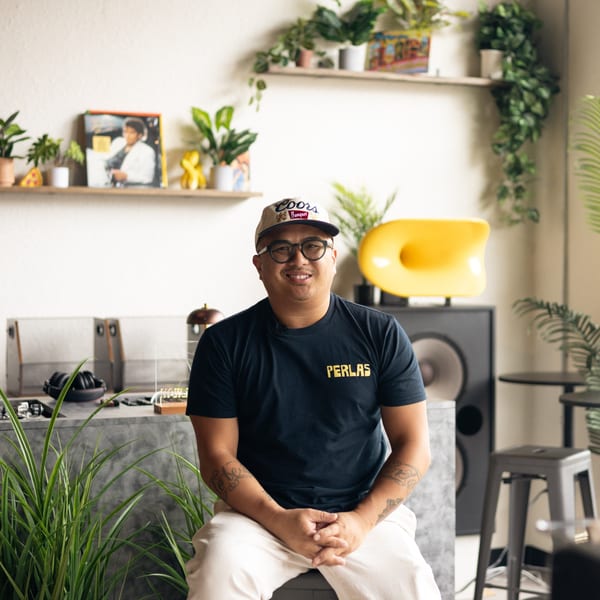 Smiling man in a Perla's Pizza shirt and Coors hat sitting in a modern, plant-filled room.